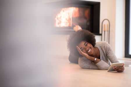 beautiful young black women using tablet computer on the floor of her luxury home in front of fireplace at autumn dayの写真素材