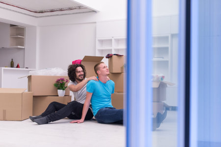 Relaxing in new house. Cheerful young gay couple sitting on the floor while cardboard boxes laying...