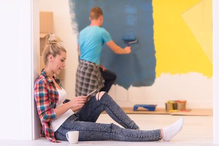 Happy couple doing home renovations, the man is painting the room and the woman is relaxing on the floor and connecting with a tabletの写真素材