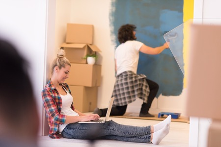 Happy couple doing home renovations, the man is painting the room and the woman is relaxing on the floor and connecting with a laptopの写真素材