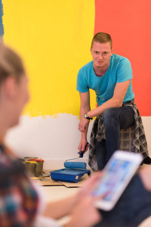 Happy couple doing home renovations, the man is painting the room and the woman is relaxing on the floor and connecting with a tabletの写真素材