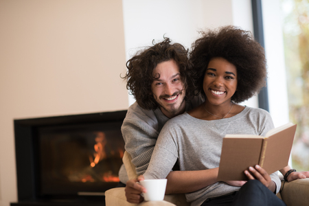 Young beautiful multiethnic couple hugging in front of fireplace at home when reading a book at autumn dayの写真素材