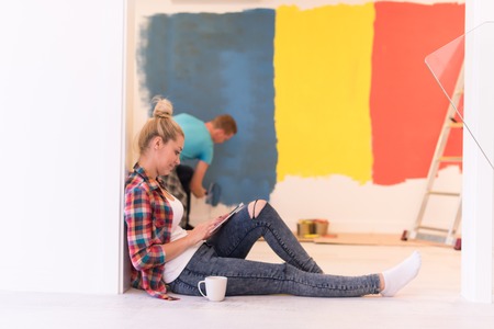 Happy couple doing home renovations, the man is painting the room and the woman is relaxing on the floor and connecting with a tabletの写真素材
