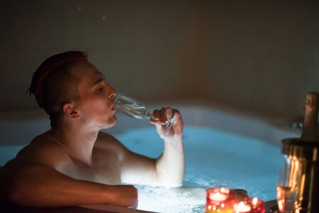 young handsome man enjoys relaxing in the bathtub with candles and champagne at luxury resort spaの写真素材