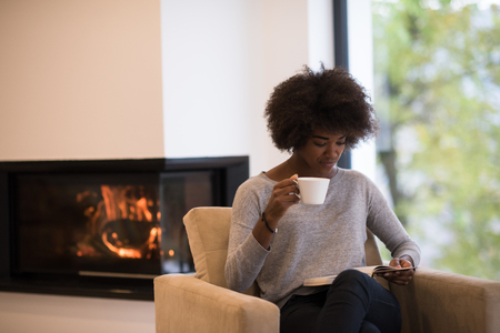 african american woman drinking cup of coffee reading book at fireplace. Young black girl with hot beverage relaxing heating warming up. autumn at home.の写真素材