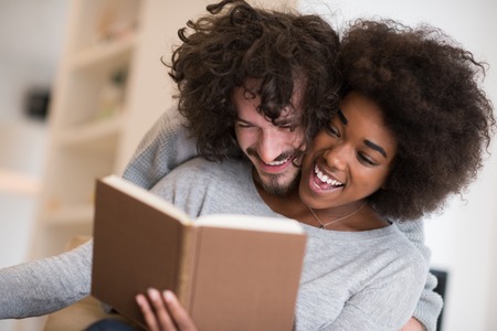 Young beautiful multiethnic couple hugging in front of fireplace at home when reading a book at autumn dayの写真素材
