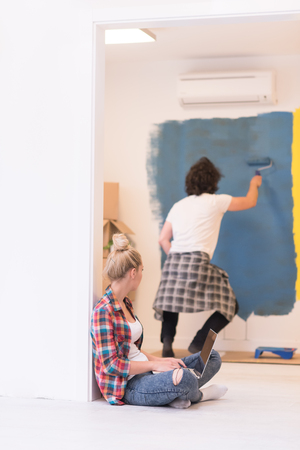 Happy couple doing home renovations, the man is painting the room and the woman is relaxing on the floor and connecting with a laptopの写真素材
