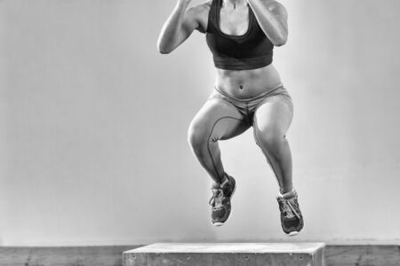 Fit young african american woman box jumping at a crossfit style gym. Female athlete is performing box jumps at gym with focus on legsの写真素材