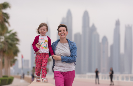 young mother and cute little girl running and cheerfully spend their time on the promenade by the sea with a big city in the backgroundの写真素材