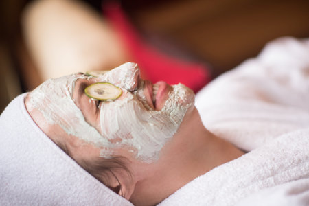 Beautiful young woman is getting facial clay mask at spa, lying with cucumbers on eyes