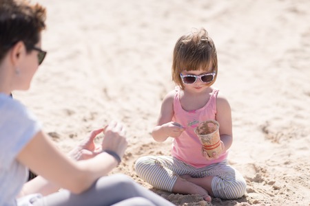 cool young mom and her little girl playing with a bucket full of sand wearing sunglasses and relaxing on the beach on a sunny dayの写真素材
