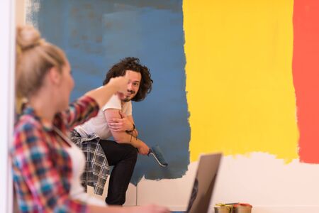 Happy couple doing home renovations, the man is painting the room and the woman is relaxing on the floor and connecting with a laptopの写真素材