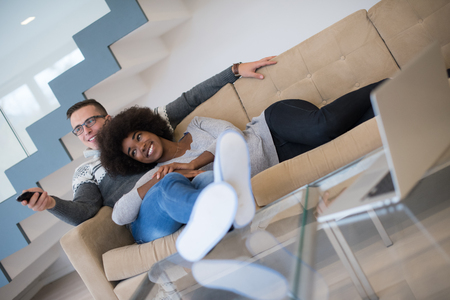 Young multiethnic couple relaxing on the sofa in the luxury living room, using a laptop and remote controlの写真素材