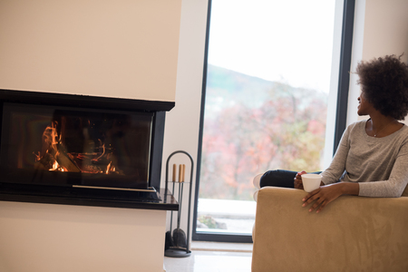 Happy joyful african american woman drinking cup of coffee relaxing at fireplace. Young black girl with hot beverage heating warming up. autumn at home.の写真素材