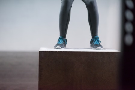 Fit young african american woman box jumping at a crossfit style gym. Female athlete is performing box jumps at gym with focus on legsの写真素材