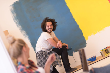Happy couple doing home renovations, the man is painting the room and the woman is relaxing on the...