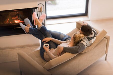Young romantic couple sitting on sofa in front of fireplace at home, looking at each other, talking and drinking coffee at autumn dayの写真素材