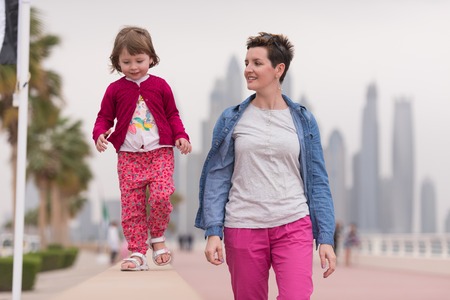 young mother and cute little girl running and cheerfully spend their time on the promenade by the sea with a big city in the backgroundの写真素材