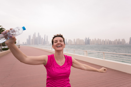 young woman celebrating a successful training run on the promenade by the sea with a bottle of water and her hands raised in the air with a big city in the backgroundの写真素材