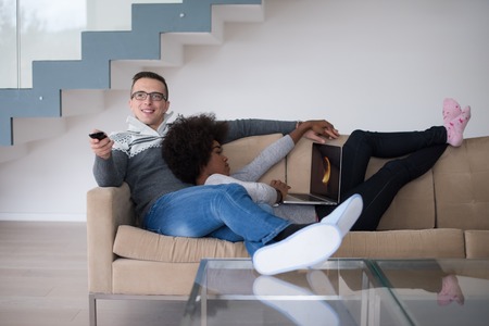Young multiethnic couple relaxing on the sofa in the luxury living room, using a laptop and remote controlの写真素材