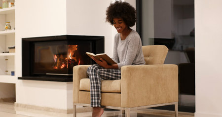 Young beautiful african american woman sitting in front of fireplace at home on a cold autumn day and reading bookの写真素材