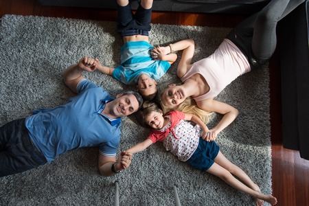 Top view of beautiful young parents, their cute little daughter and son holding hands, looking at camera and smiling, lying on the floorの写真素材