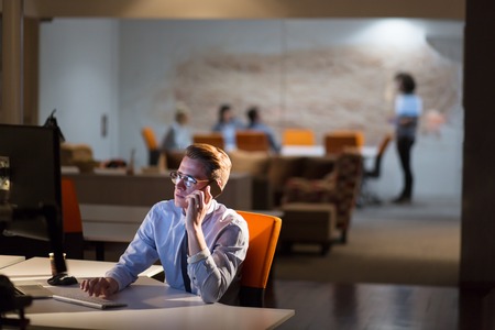 Young man using mobile phone while working on computer at night in dark office.の写真素材