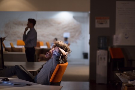 Full length of a relaxed casual young businessman sitting with legs on desk at night officeの写真素材