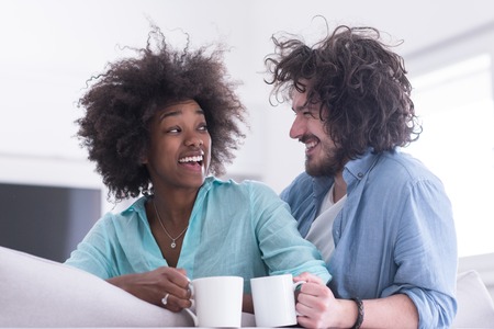 young multiethnic couple sitting on sofa at home drinking coffe, talking, smiling.の写真素材