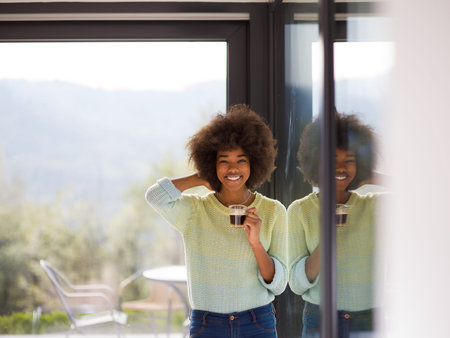 Beautiful young african american woman drinking coffee and looking through a window in her luxury...