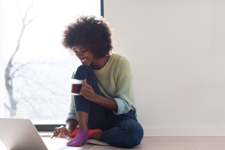Young african american woman smiling sitting on the floor near bright window while looking at open laptop computer and holding mug at homeの写真素材