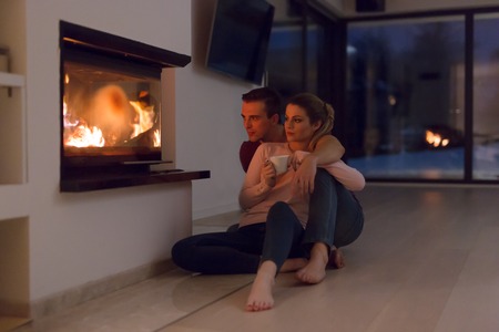 Young romantic couple sitting on the floor in front of fireplace,talking and drinking tea on cold winter night at homeの写真素材
