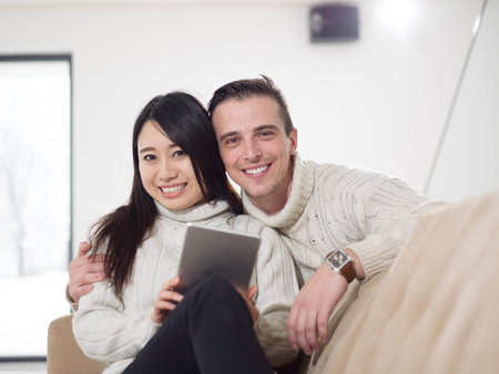 Young multiethnic couple relaxing at luxury home with tablet computers reading in the living room on the sofa couch.の写真素材
