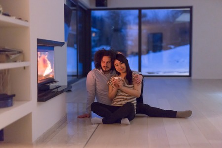 Young romantic multiethnic couple sitting on the floor in front of fireplace at home, talking and drinking tea on cold winter nightの写真素材