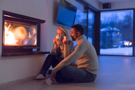 Young romantic couple sitting on the floor in front of fireplace,talking and drinking tea on cold winter night at homeの写真素材