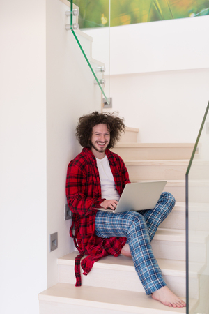 Young freelancer in bathrobe working from home using laptop computer while sitting on stairsの写真素材