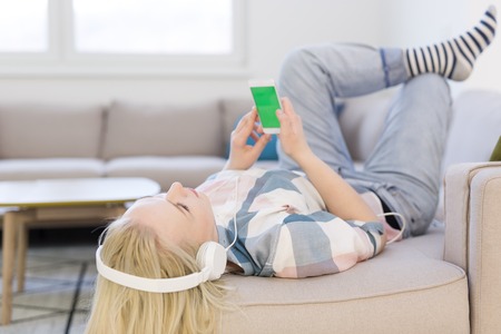 Young beautiful girl enjoying music through headphones, laying on sofa at homeの写真素材