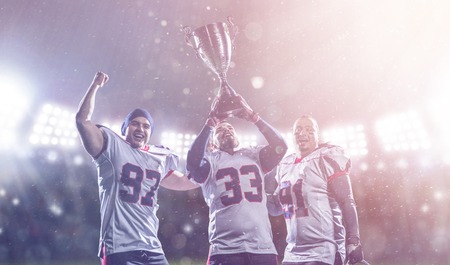 happy american football team with trophy celebrating victory in the cup final on big modern stadium with lights and flares at night