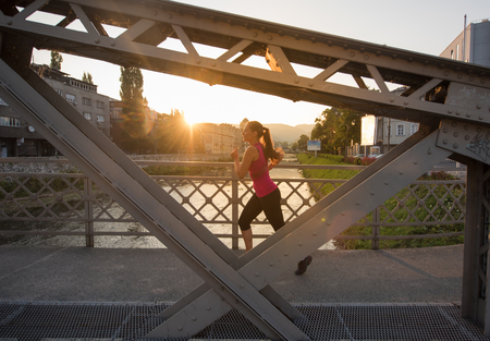 young sporty woman jogging across the bridge at sunny morning in the cityの写真素材