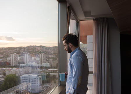 young man enjoying evening coffee and beautiful sunset landscape of the city while standing by the windowの写真素材