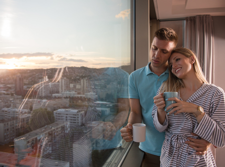 romantic happy young couple enjoying evening coffee and beautiful sunset landscape of the city while standing by the windowの写真素材