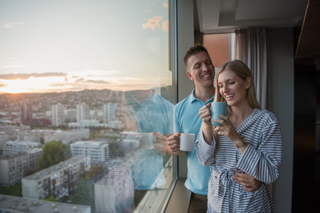 romantic happy young couple enjoying evening coffee and beautiful sunset landscape of the city while standing by the windowの写真素材