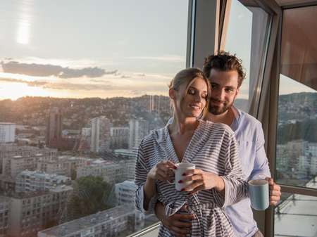 romantic happy young couple enjoying evening coffee and beautiful sunset landscape of the city while standing by the windowの写真素材