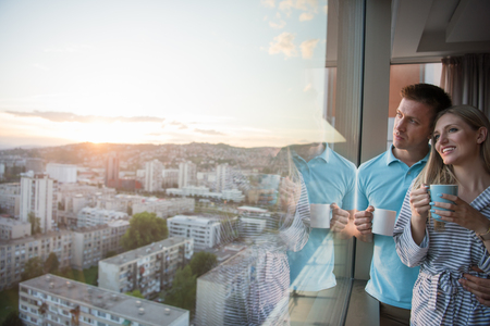 romantic happy young couple enjoying evening coffee and beautiful sunset landscape of the city while standing by the windowの写真素材