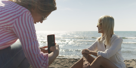 Two Girl Friends Taking Photo with Smartphone On Empty Beach during autumn dayの写真素材