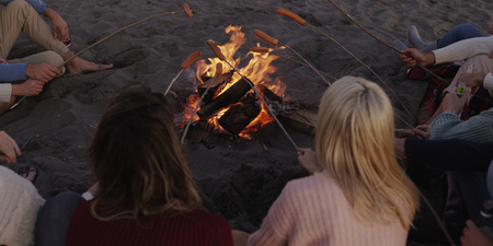 Group of young friends sitting by the fire late at night, grilling sausages and drinking beer, talking and having funの写真素材