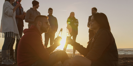 Happy Carefree Young Friends Having Fun And Drinking Beer By Bonefire On The Beach As The Sun Begins To Setの写真素材