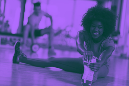 happy young african american woman in a gym stretching and warming up before workout young mab exercising with dumbbells in background duo tone filterの写真素材
