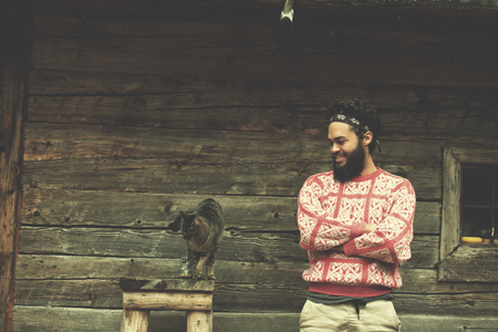 portrait of young hipster man with beard and cat in front of wooden houseの写真素材