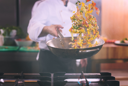 Young male chef flipping vegetables in wok at commercial kitchenの写真素材